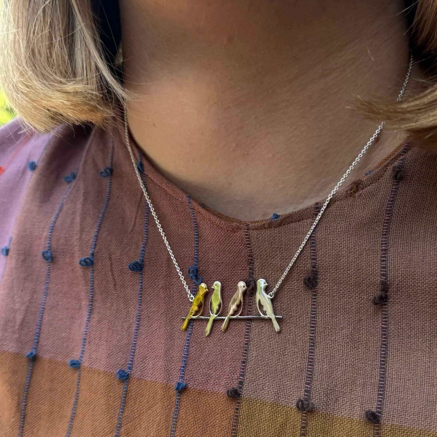 A sterling silver supersize bird necklace, with four bird silhouettes in silver, gold and rose gold plate and sat on a branch. The design hangs centrally on a fine silver chain and worn on a models neck.