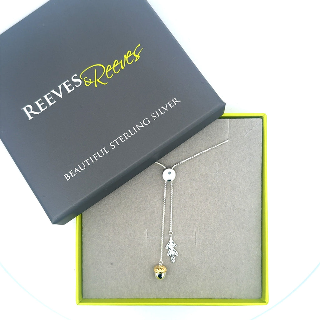 An Acorn and Oak Leaf Charm hanging from fine silver chains and threaded through a silver slider clasp. Shown in a grey and green Reeves & Reeves branded gift box.