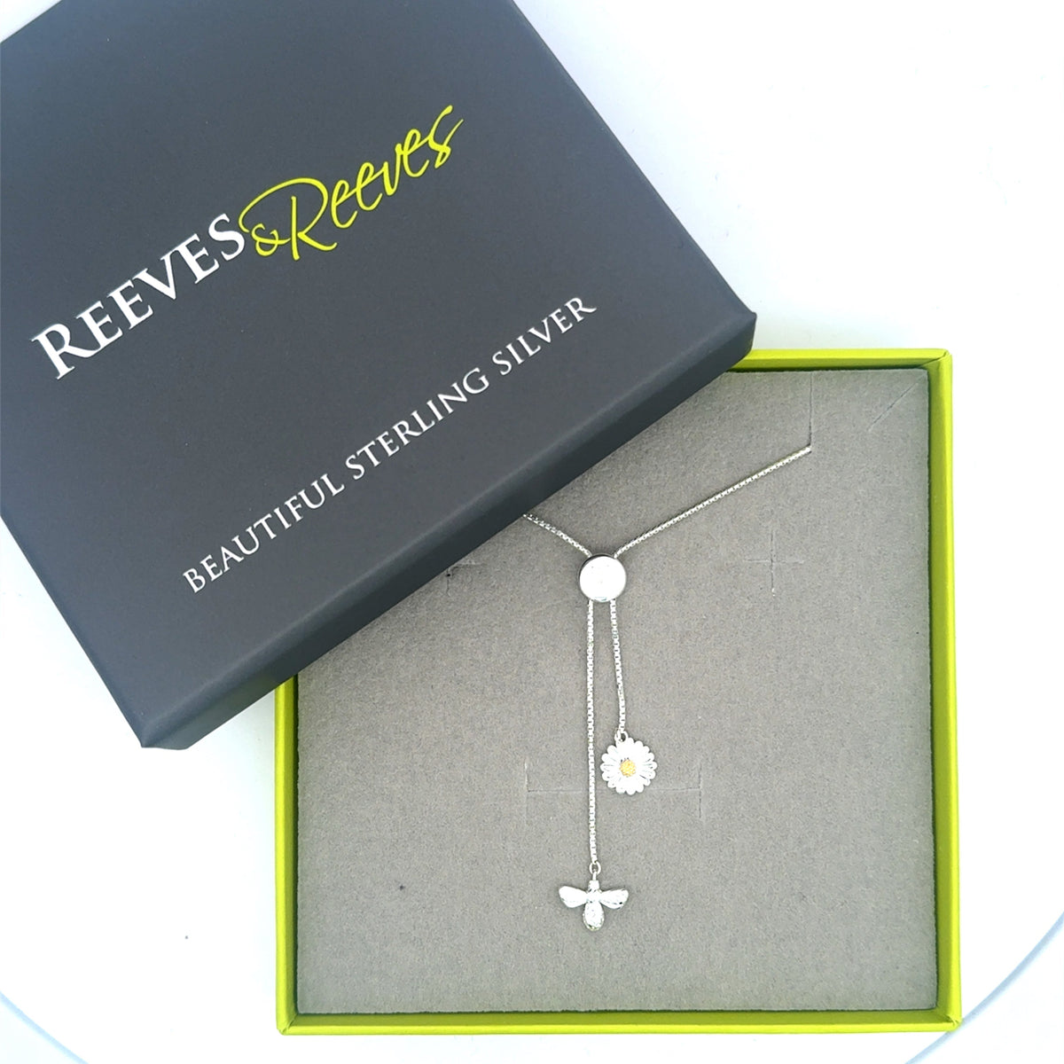 A fine silver slider chain necklace with a daisy and bee charm hanging from the slider toggle. Shown in a Reeves & Reeves grey and green gift box.