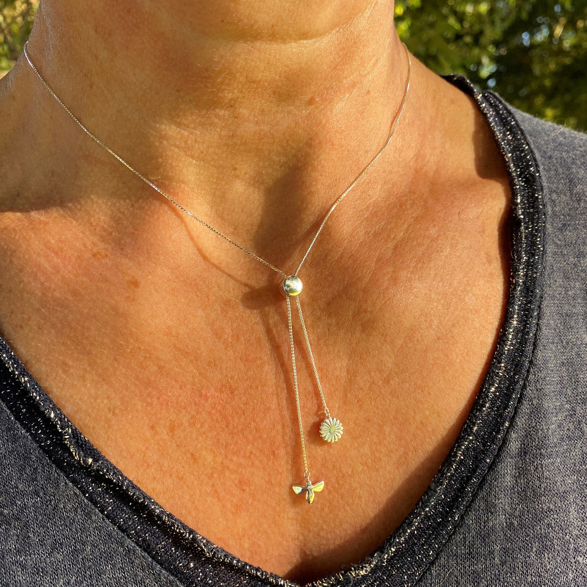 A silver necklace with a daisy and a bee being worn on a model with a blue jumper