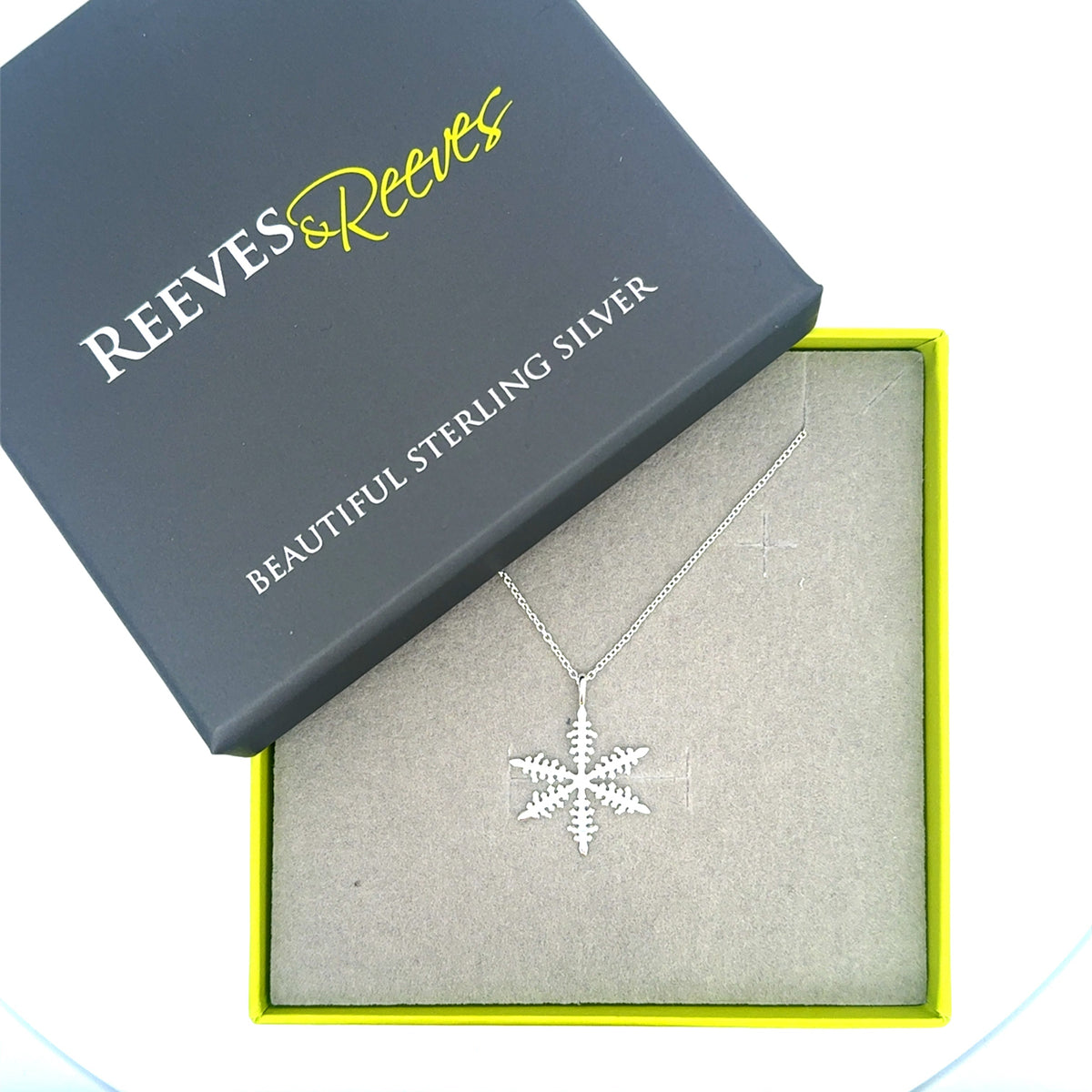 A sterling silver snowflake pendant, hanging on a silver chain and shown in a Reeves & Reeves grey and green gift box.