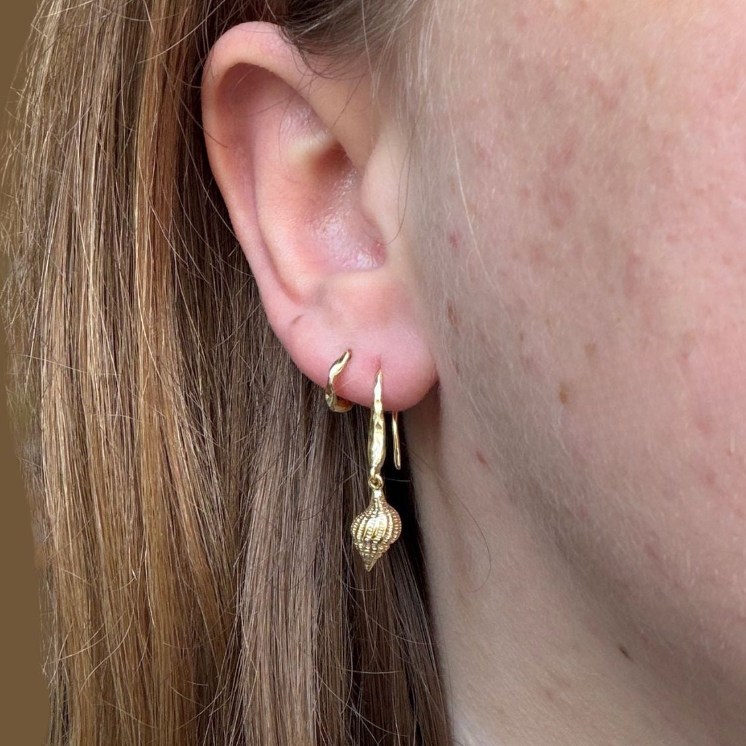 Gold plated conch shell drop earrings worn in a ladies ear with a smaller gold plate hoop in her second piercing.
