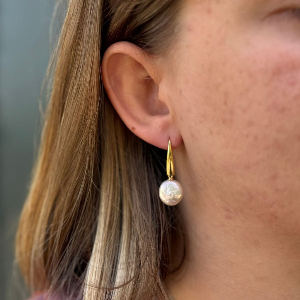 Gold plated hook earrings with a white button pearl hanging from the bottom of the hook, worn in a ladies ear.