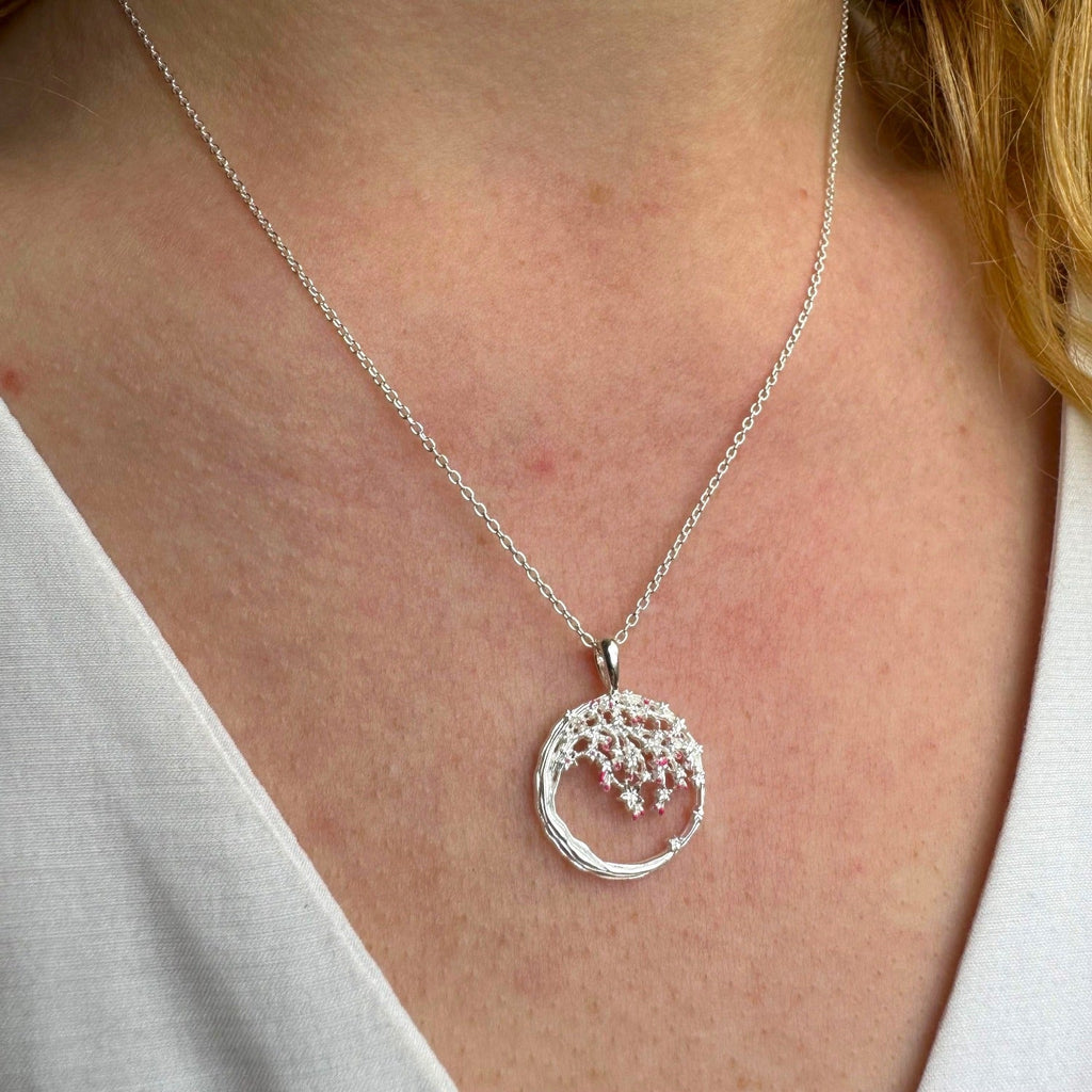 A Silver circular pendant with a cherry blossom design sat inside as if dangling from a tree. The design is finished with pink enamel detail and hung on a silver chain which is worn on a models neck.