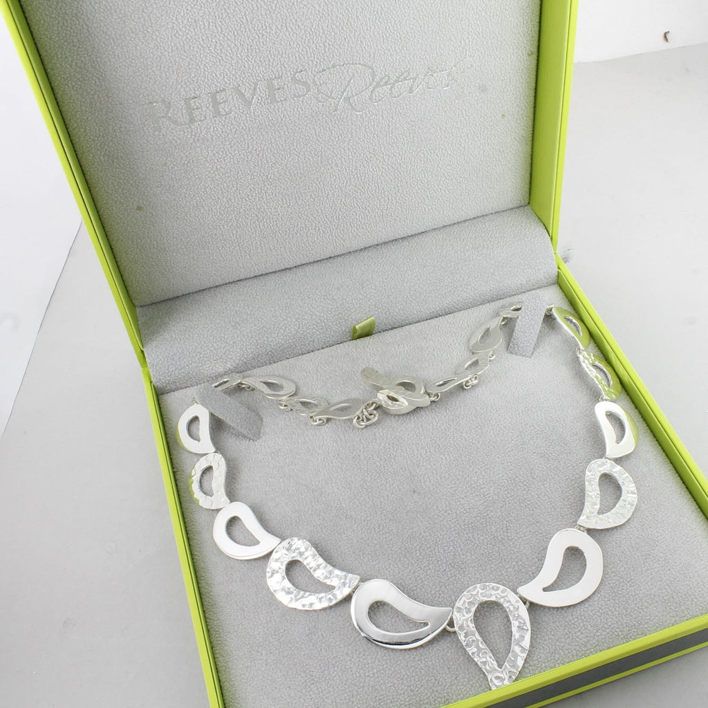 A sterling silver necklace with droplet shapes in both high shine and hammered finishes, shown in a Reeves & Reeves grey and green gift box.