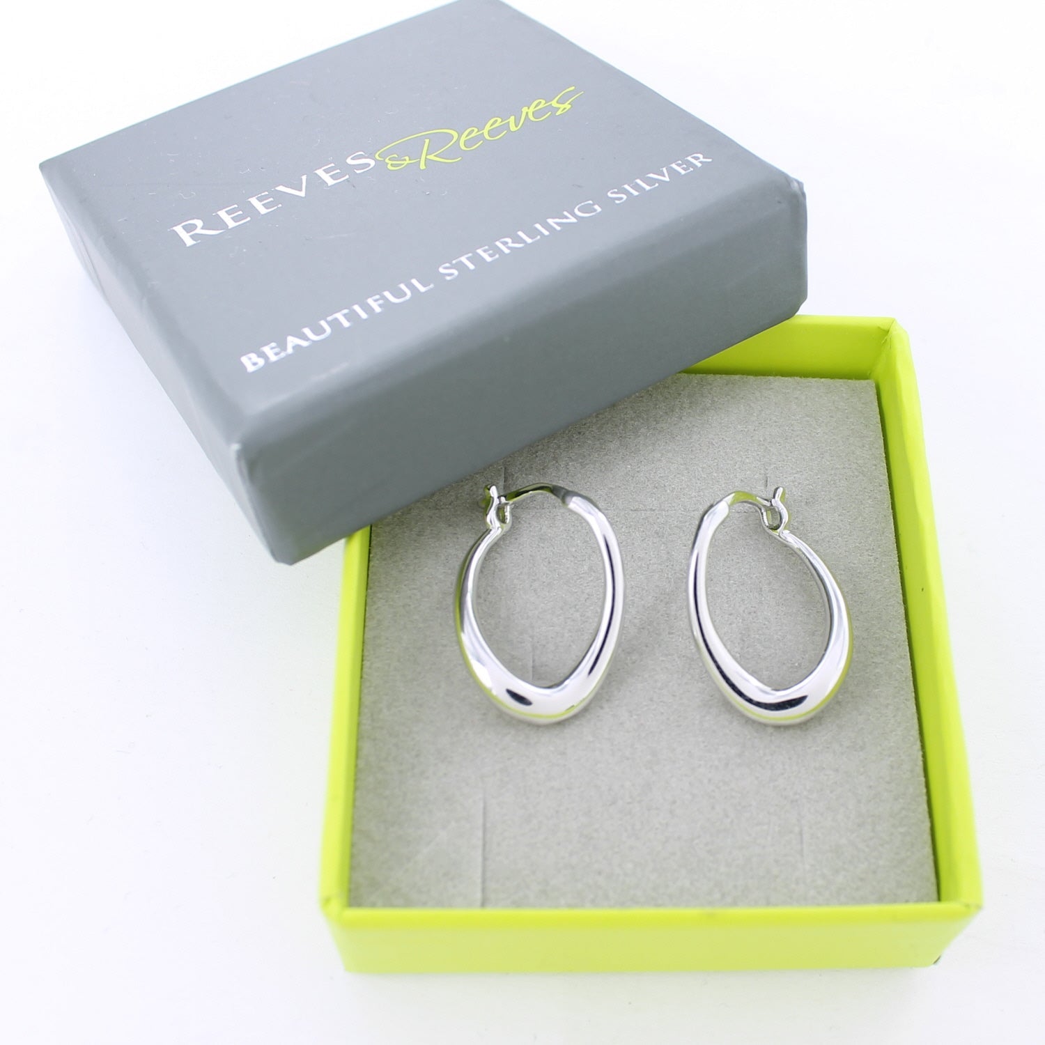A pair of silver hoop earrings with a hinged clasp, shown in a grey and green Reeves & Reeves branded gift box.