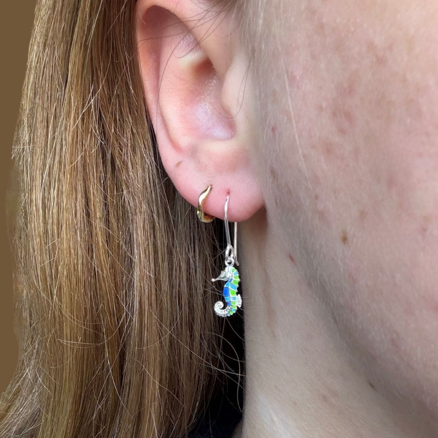 Sterling Silver Seahorse Earrings with blue and green enamel detail hanging on silver hooks and worn in a ladies ear with a small gold hoop earring.