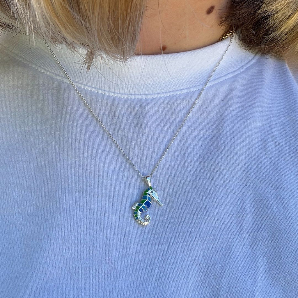A seahorse pendant with blue and green enamel detail worn by a person on a white t shirt.