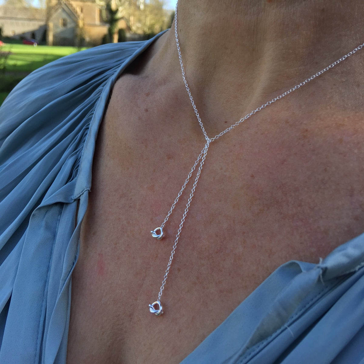 A close-up of a person wearing a delicate silver Y-necklace with two small openwork knot pendants, styled with a blue draped top.