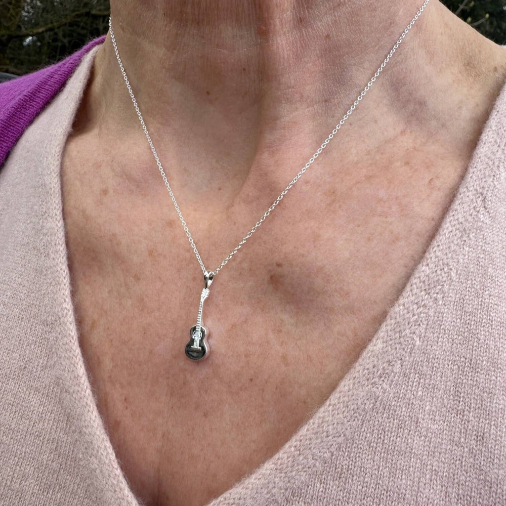 A Sterling Silver pendant in the shape of a guitar, hanging on a silver chain and worn on a models neck.