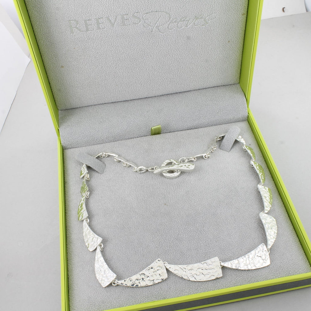 A sterling silver necklace comprised of sail style shapes that have a hammered finish, shown in a Reeves & Reeves grey and green gift box.