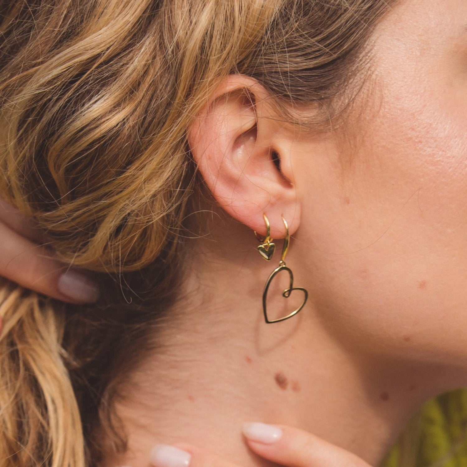 A sterling silver hoop earring with gold plate finish and a heart charm hanging from the bottom. Shown in a ladies ear with a larger looped heart drop earring.