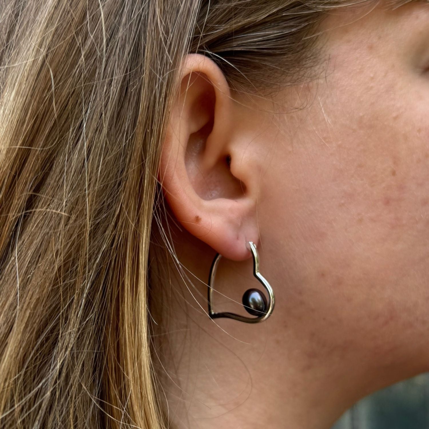 Silver heart shaped hoop earring with a peacock colour pearl sat inside the heart silhouette. Worn in a ladies ear.
