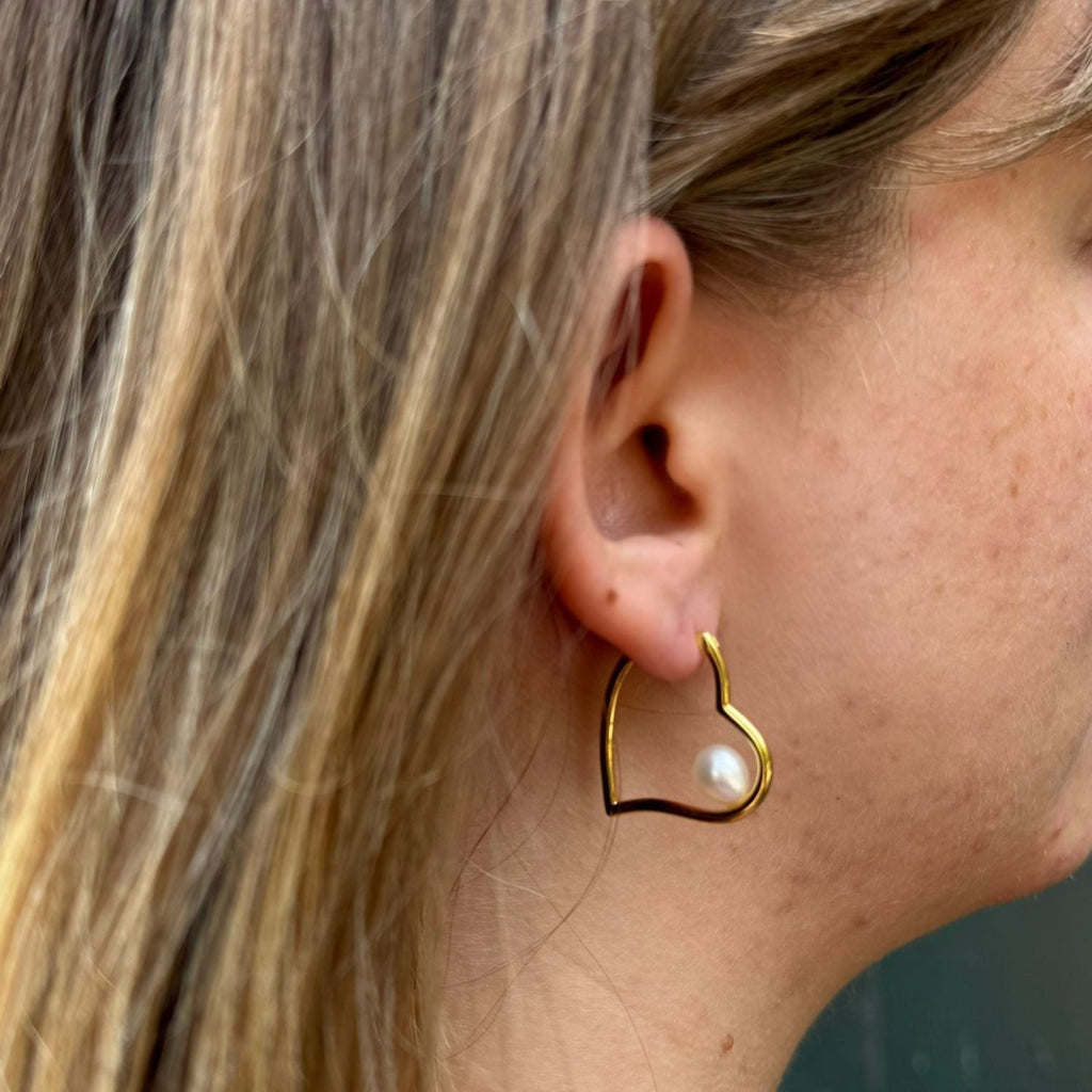 Gold plated heart-shaped hoop earring with a white freshwater pearl sat inside, worn on a woman's ear.