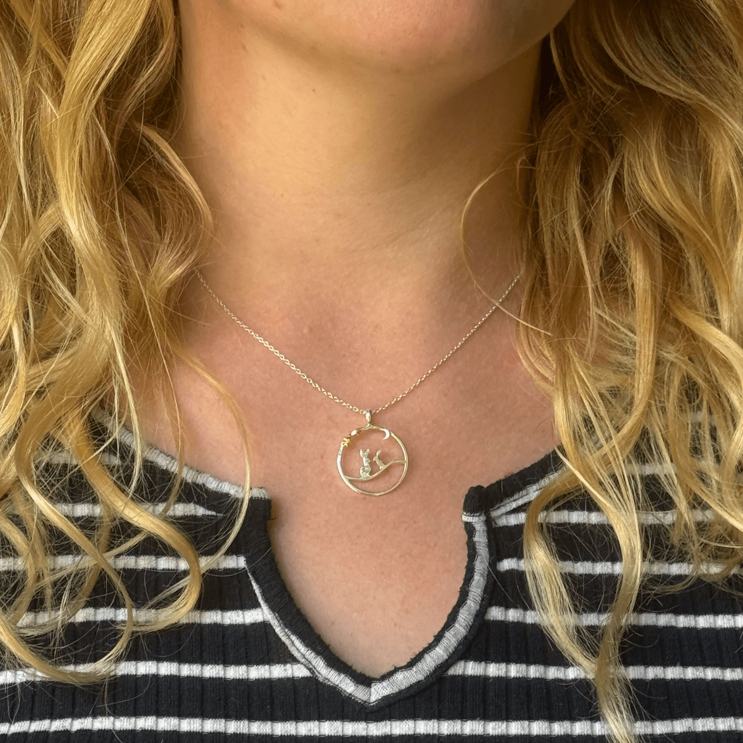 Two Silver Hares sat in a circle outline underneath a silver moon and two gold plate stars. The pendant sits on an adjustable chain and is worn on a model. Nature inspired gift for her.