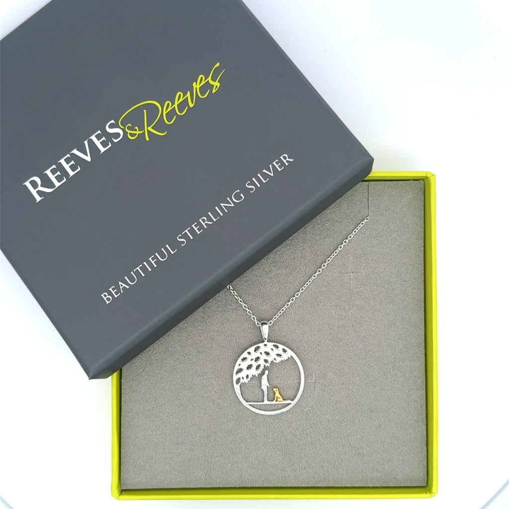 A sterling silver circular pendant with a silhouette of a person and an 18ct gold plated dog sat under the tree. Hung on a silver chain and shown in a Reeves & Reeves gift box.