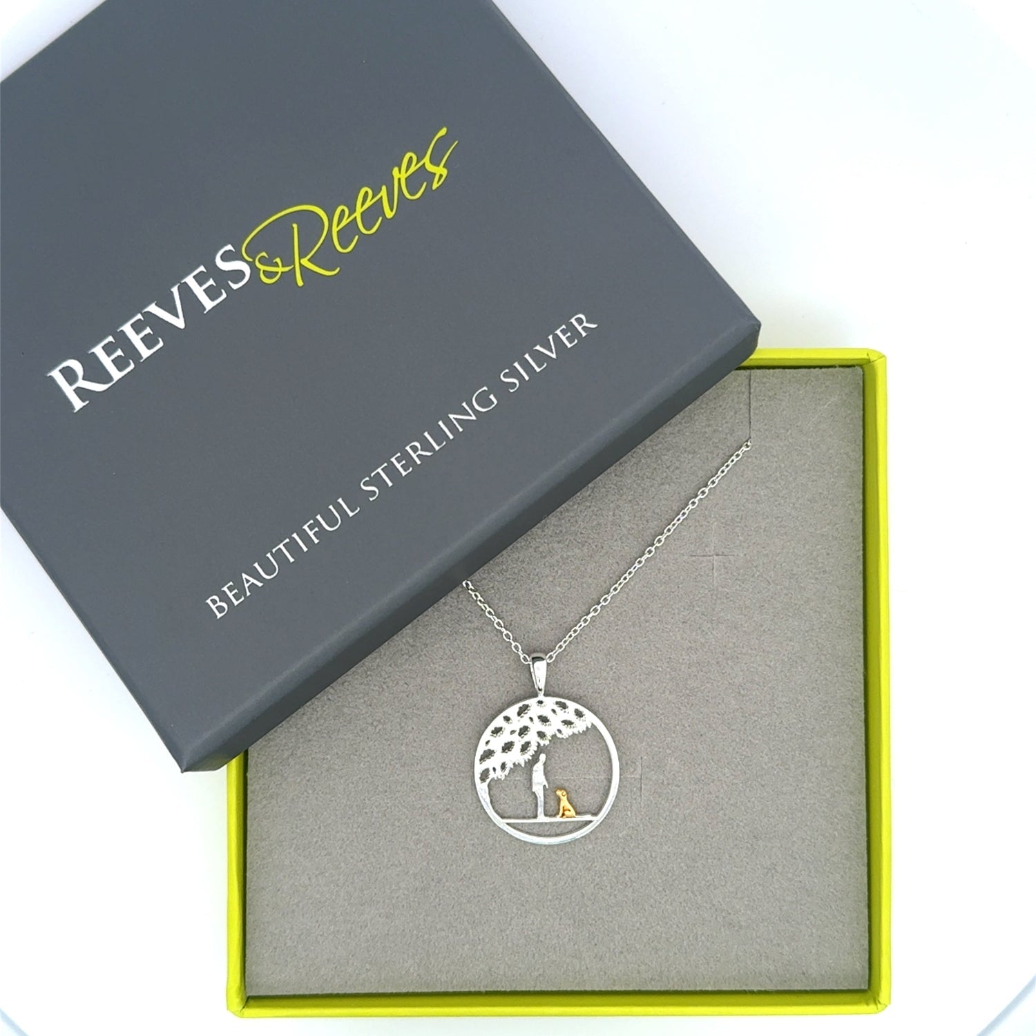 A sterling silver circular pendant with a silhouette of a person and an 18ct gold plated dog sat under the tree. Hung on a silver chain and shown in a Reeves & Reeves gift box.
