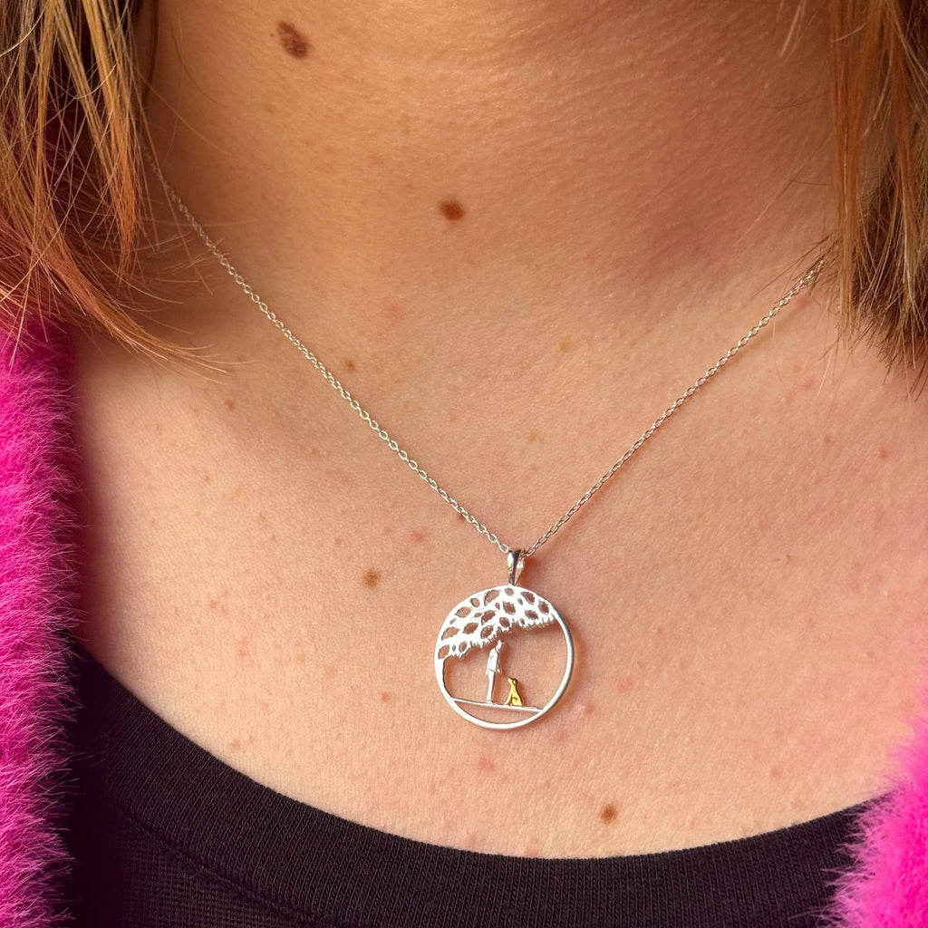 A sterling silver circular pendant with a tree design inside, and a person silhouette with an 18ct gold plated dog sat under the tree. Hung on a silver adjustable chain and worn on a model.