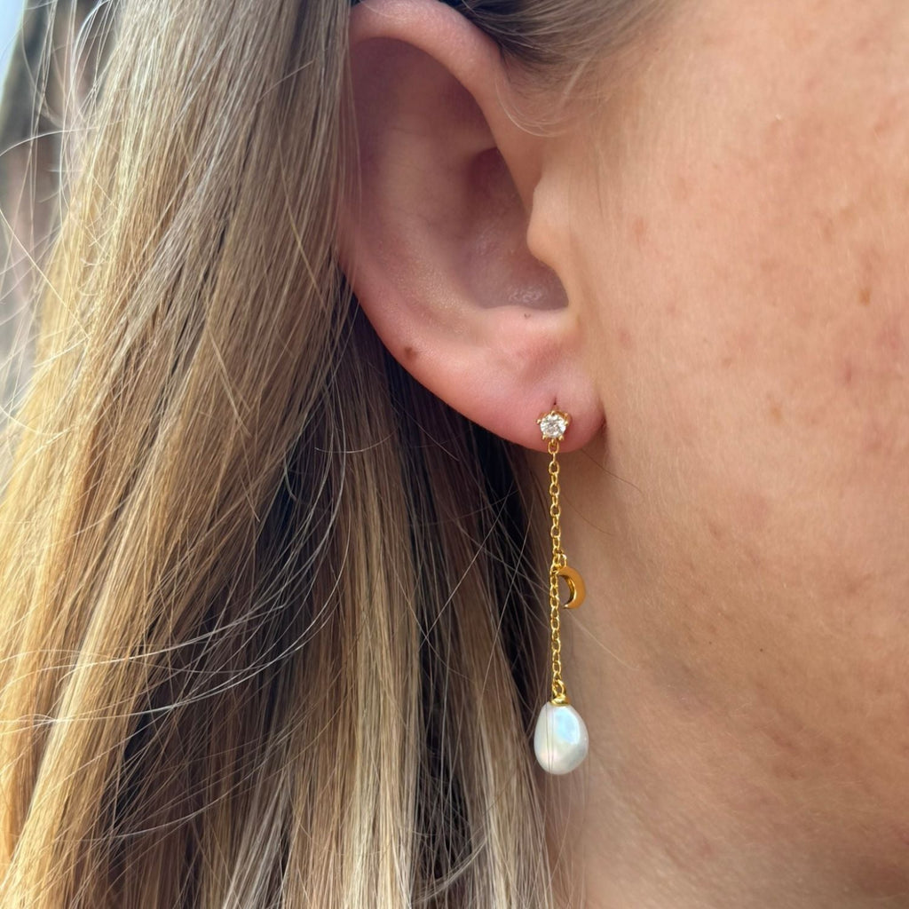 Gold plate drop earrings with a cubic zirconia stud and a chain hanging below with a moon charm halfway down the chain and a freshwater white pearl at the bottom. Worn in a ladies ear.