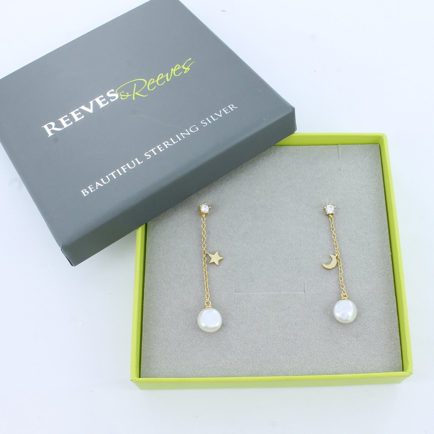 A pair of gold plate drop earrings with cubic zirconia stones, moon and star charms hanging halfway down the chain and white pearls at the bottom. Shown in a Reeves & Reeves gift box.