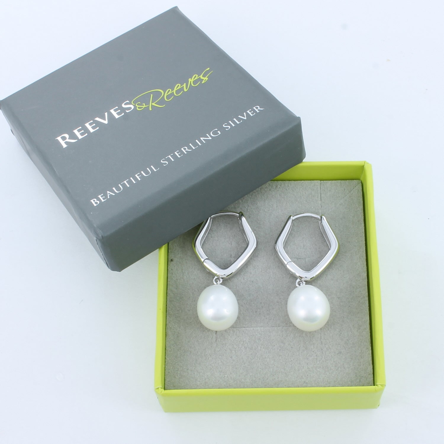 A pair of silver pentagon shaped hoop earrings with a white pearl charm. Shown in a Reeves & Reeves grey and green branded gift box.