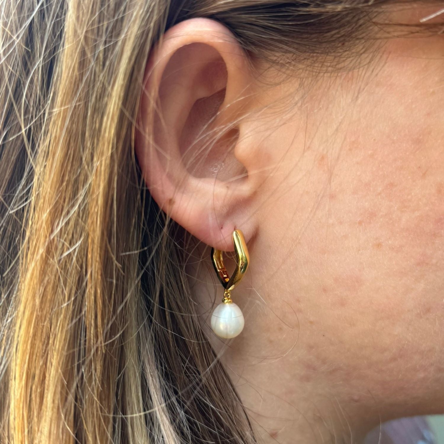 A pair of gold plated, pentagon shaped hoop earrings with a white freshwater pearl hanging from the bottom. Worn on a ladies ear.