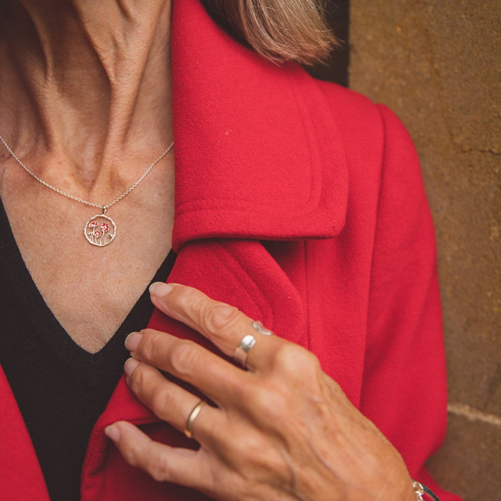 A sterling silver circular pendant with undulated edges with a poppy design finished with red enamel detail in the centre. Hung on a silver chain and shown on a models neck.