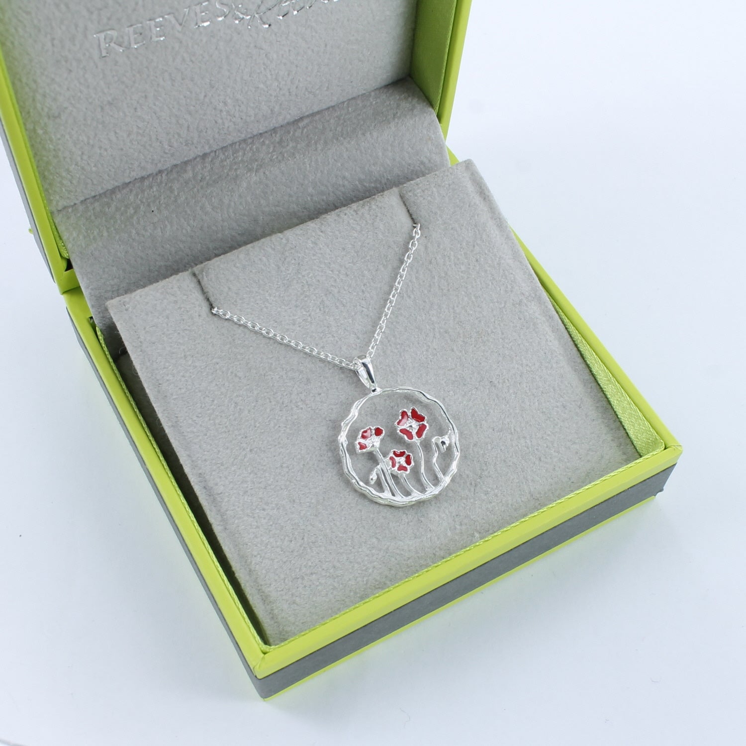 A sterling silver circular pendant with undulated edges and a poppy design in the centre. Shown in a Reeves & Reeves grey and green branded gift box.