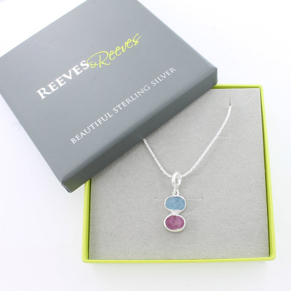 A sterling silver rough cut ruby and aqua stone sit vertically and hang on a silver chain. Shown in a grey and green Reeves & Reeves gift box.
