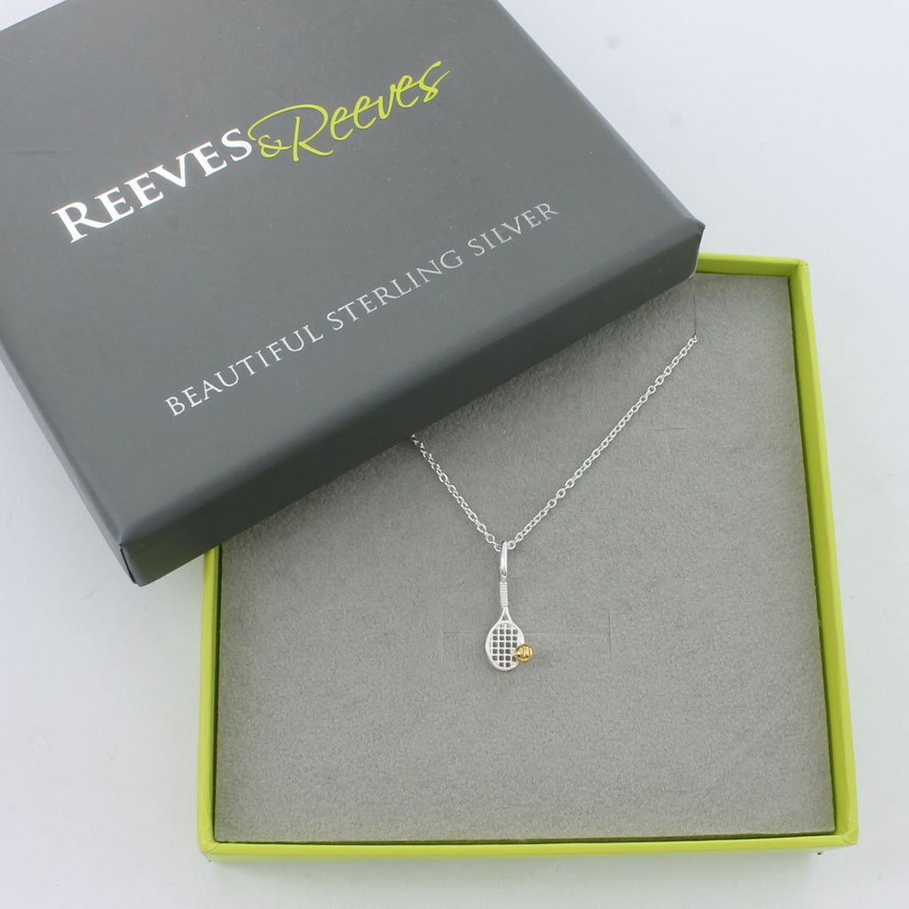 A sterling silver Tennis racket necklace with a gold plated ball sat on the edge of the racket. The pendant hangs on a silver chain and is shown in a Reeves & Reeves gift box.