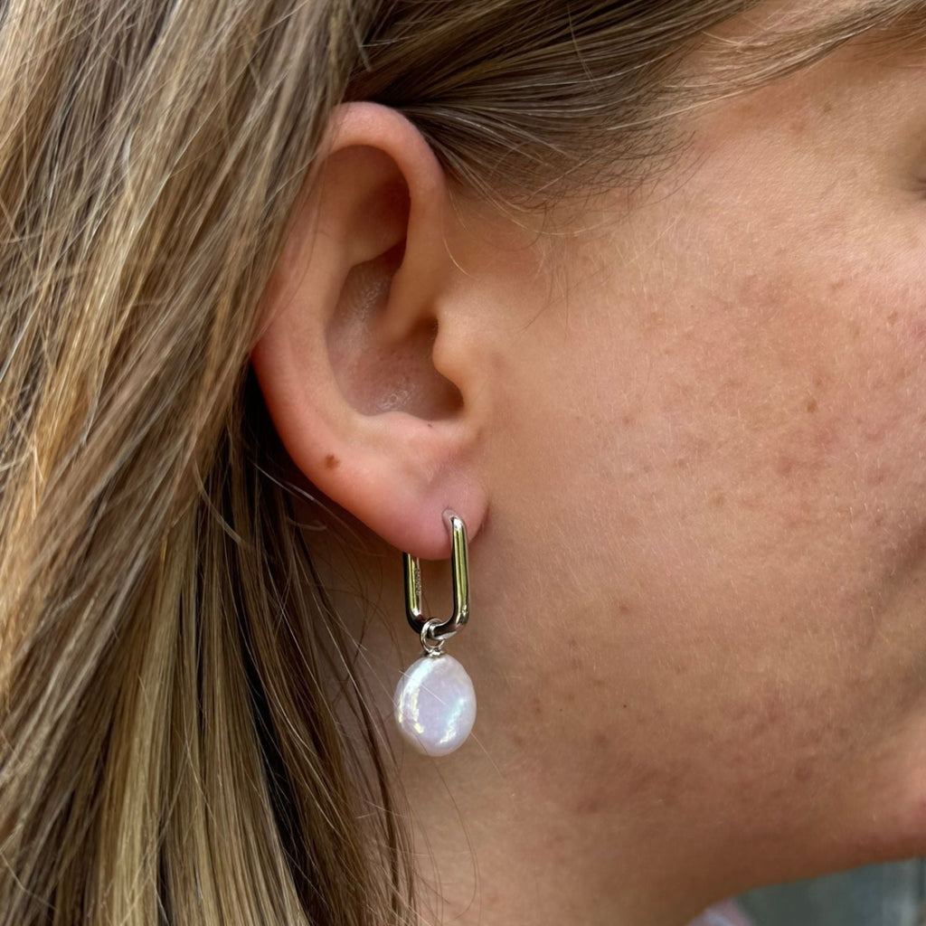 A pair of silver rectangular hoop earrings with a white button pearl charm hanging from the hoop and worn in a ladies ear.