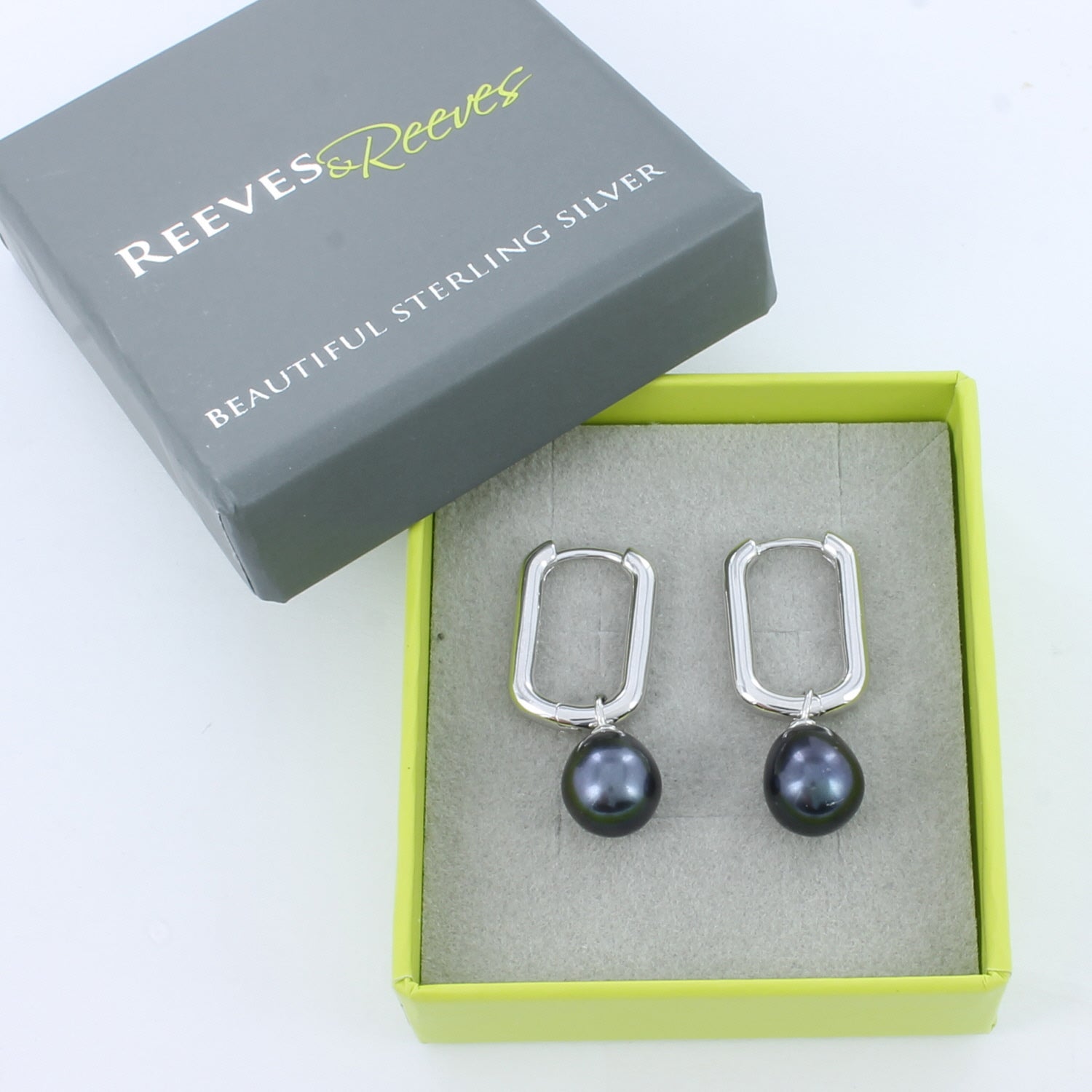 Pair of silver huggie earrings with peacock pearl charms, shown in a Reeves & Reeves grey and green branded gift box.