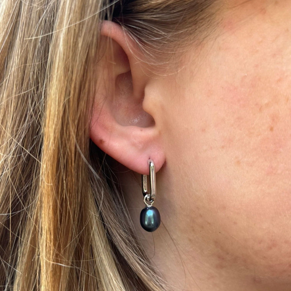 Silver rectangular huggie earrings with a peacock pearl charm hanging from the bottom, worn in a ladies ear.