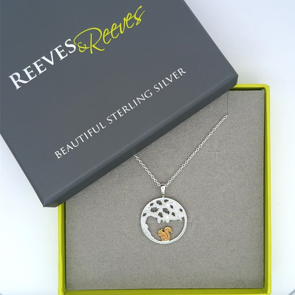 A sterling silver circular pendant with an oak tree design and a gold plated squirrel inside. Hanging on a silver adjustable chain and shown in a Reeves & Reeves grey and green gift box.