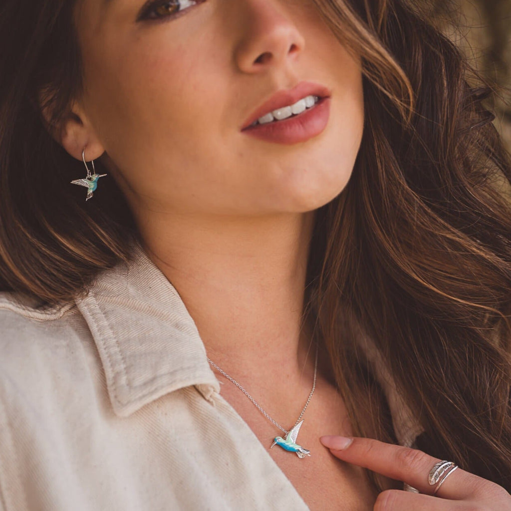 A sterling silver Hummingbird pendant with blue and green enamel detail, hanging on a silver chain and worn on a ladies neck with the matching earrings.
