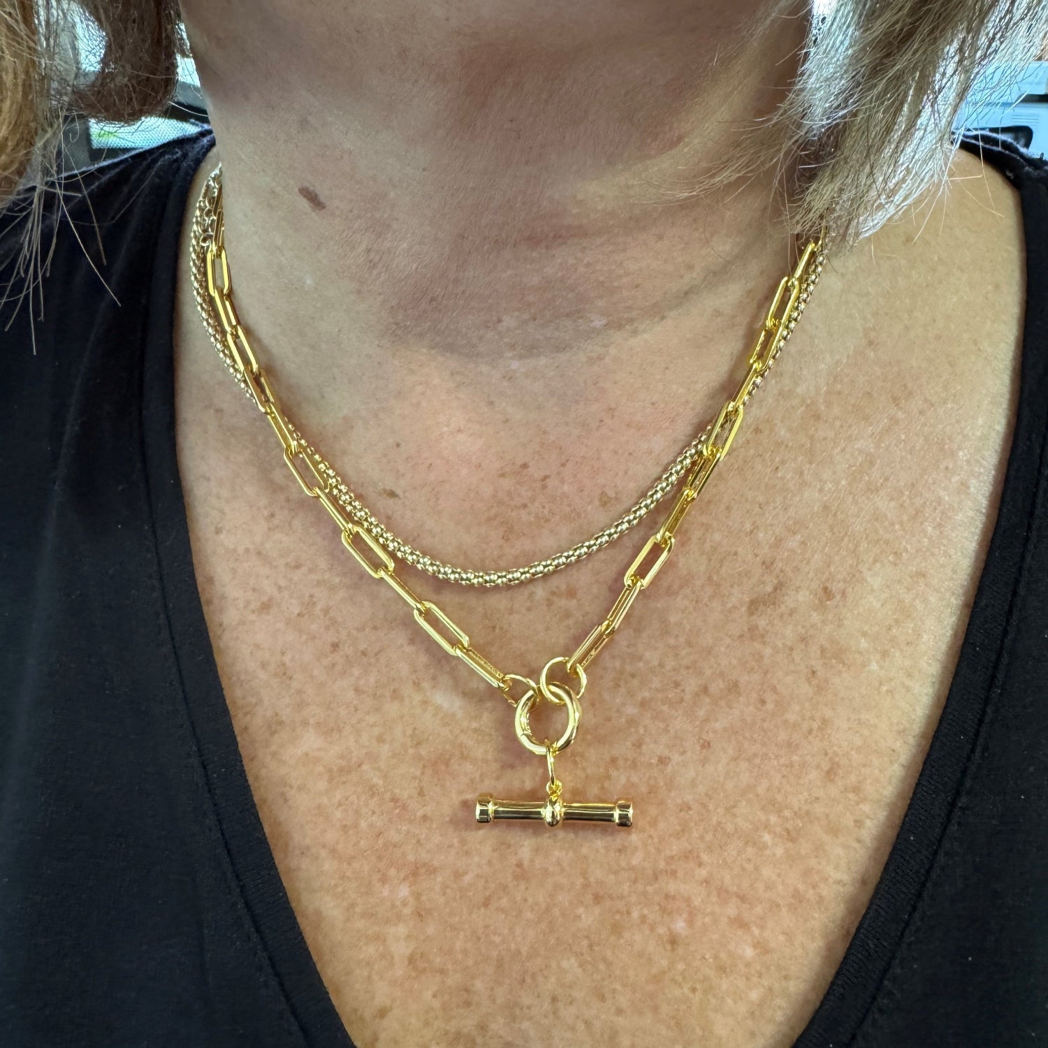 gold plated paperclip style chain necklace with a circular clasp that doubles as the central feature. A gold plated T Bar hangs from the clasp. Worn on a models neck and layered with a gold bobble chain.