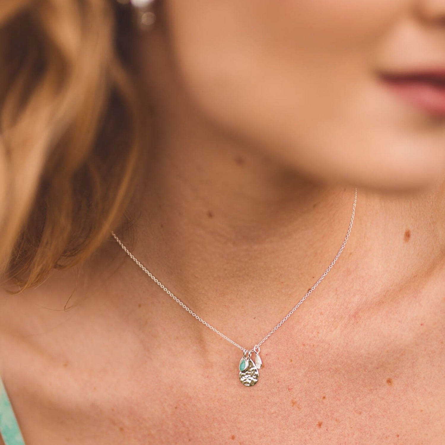 Sterling Silver Pear drop hammered charm with three smaller charms in silver and one with a turquoise stone, all hanging from a silver chain, worn on a model.