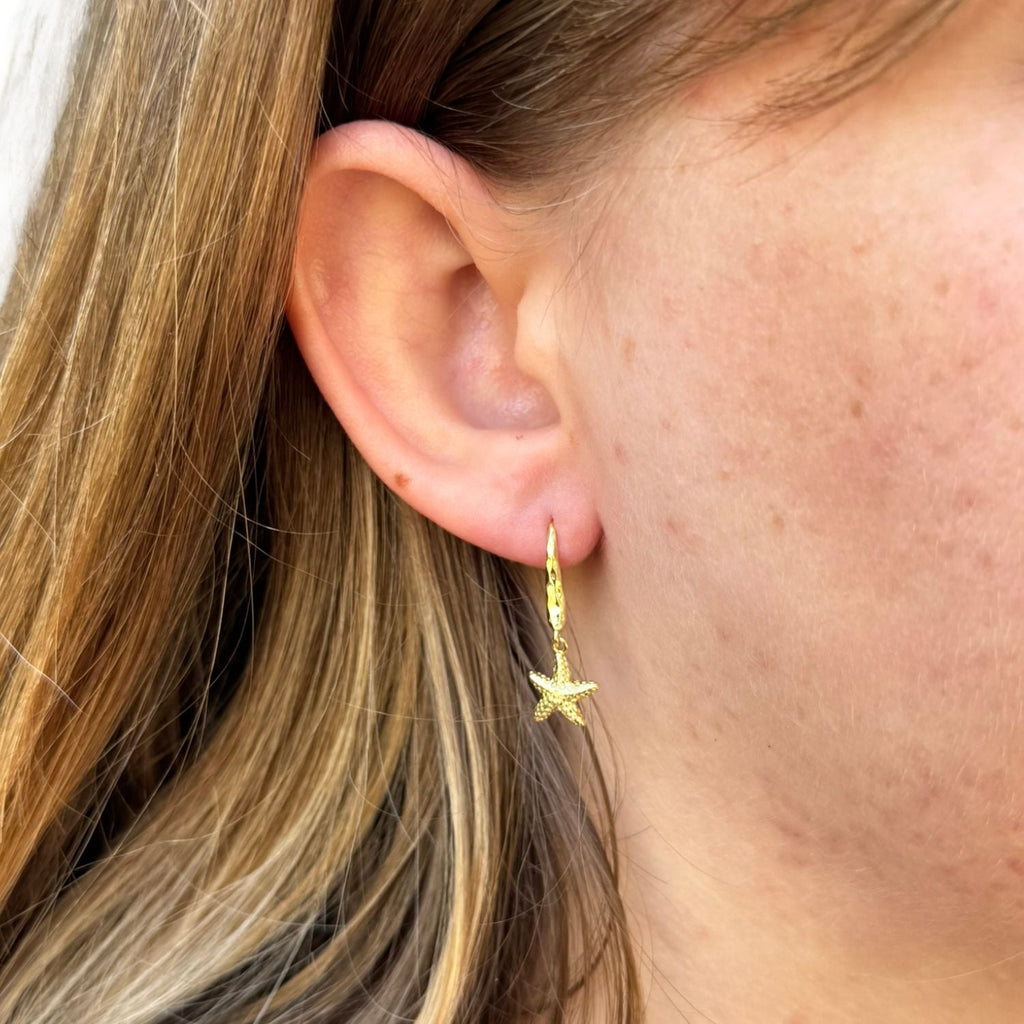 A gold plated drop earring with a detailed starfish hanging from the bottom of a hammered hook and shown in a ladies ear.