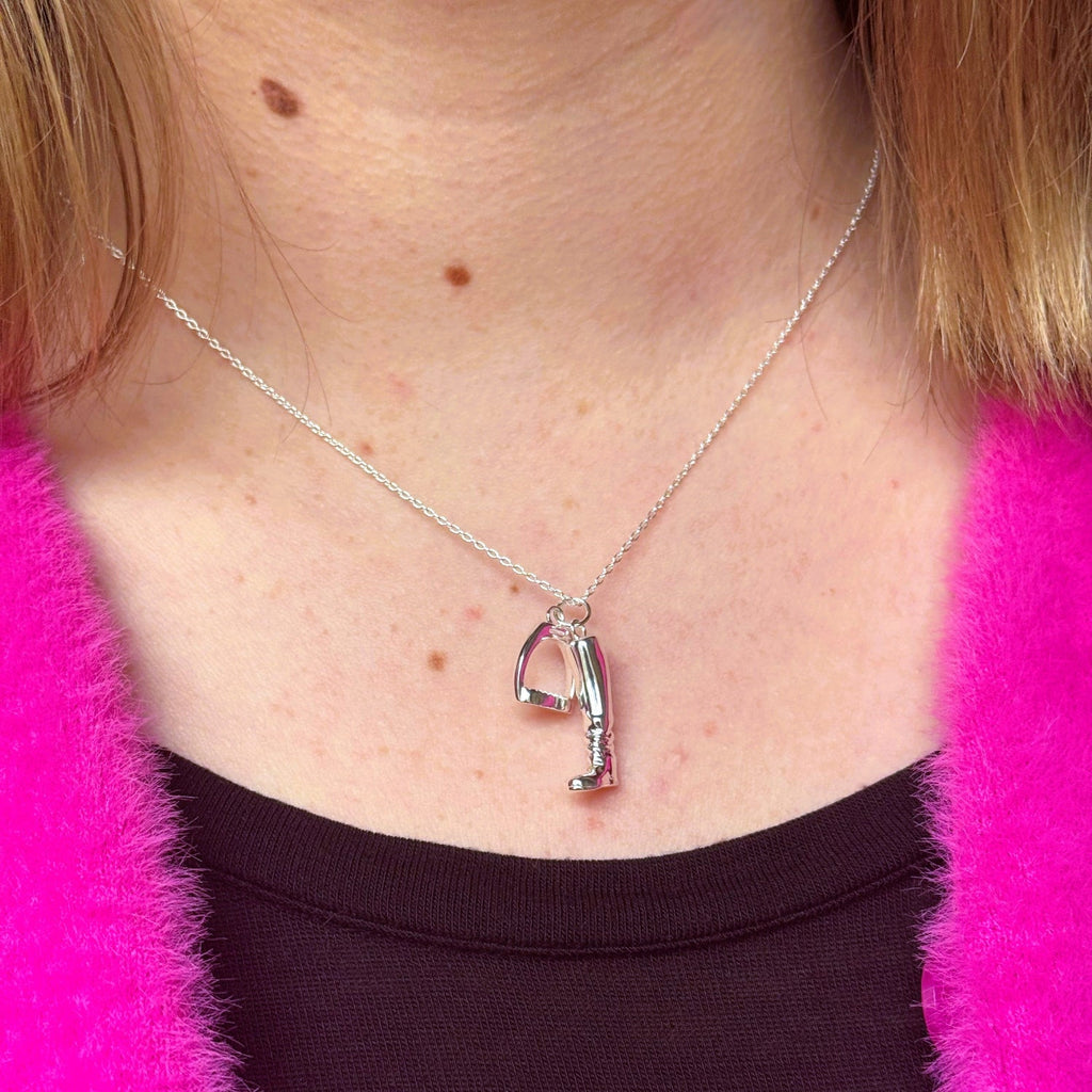 A sterling silver boot charm with a stirrup hanging next to it, both hung on a silver chain and worn on a models neck.