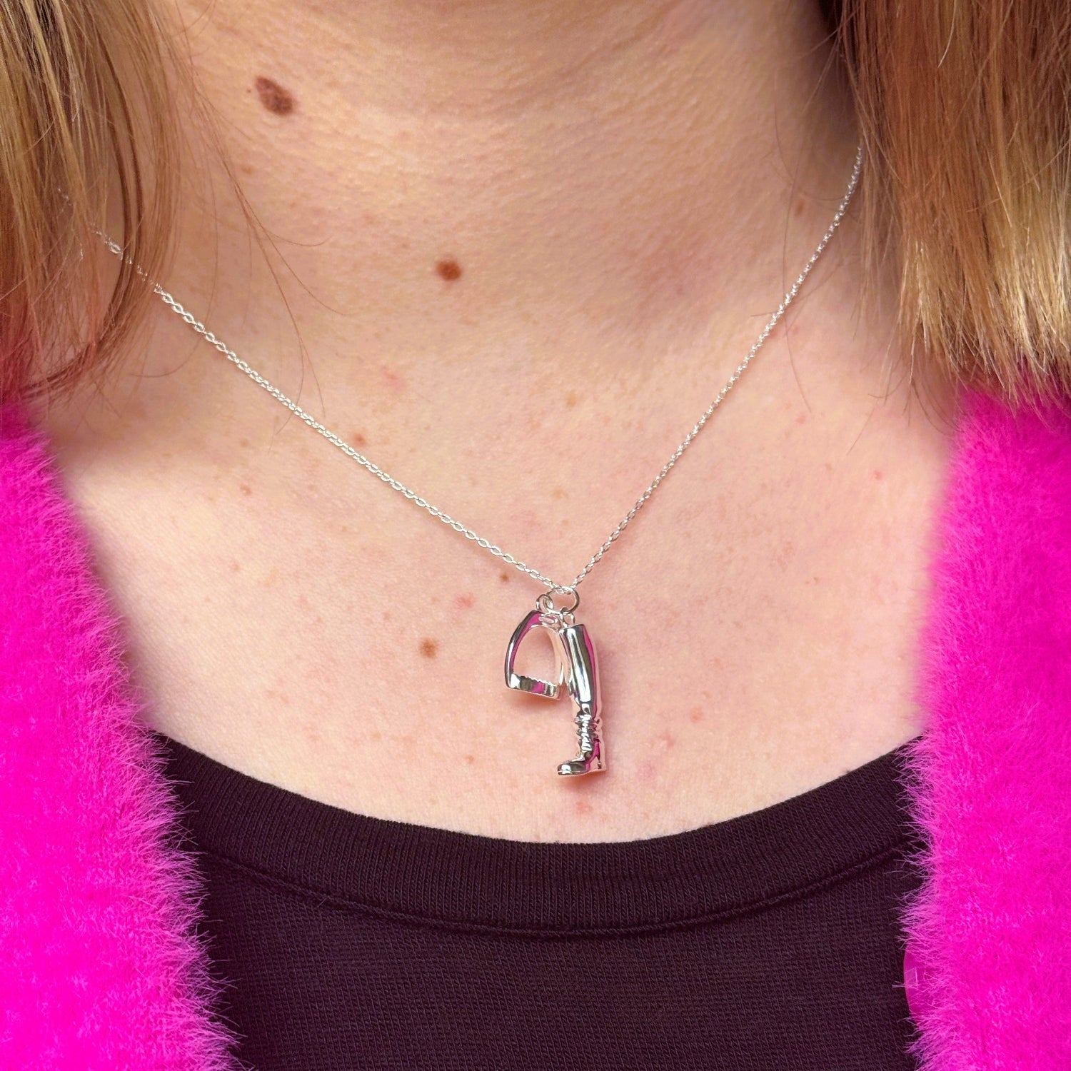 A sterling silver boot charm with a stirrup hanging next to it, both hung on a silver chain and worn on a models neck.