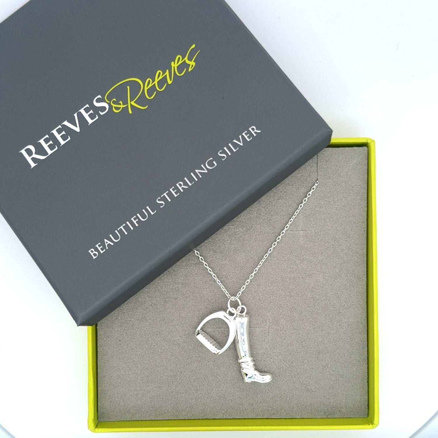 A sterling silver boot charm with a stirrup charm hanging next to it, hung on a silver chain and shown in a Reeves & Reeves grey and green gift box.