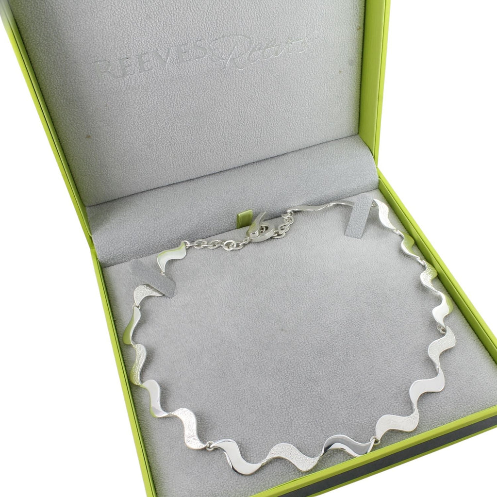 A sterling silver wave necklace, with high shine and hammered textures on the waves, finished with a t bar clasp and shown in a Reeves & Reeves gift box.