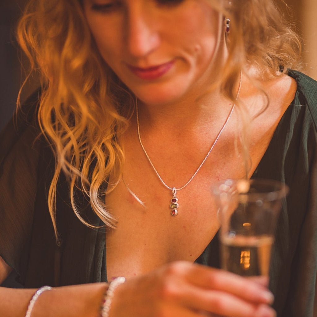A three stone pendant, with two circular ruby stones and a square peridot stone sat in the middle. Hanging from a silver rope style chain and shown on a model with the matching earrings.
