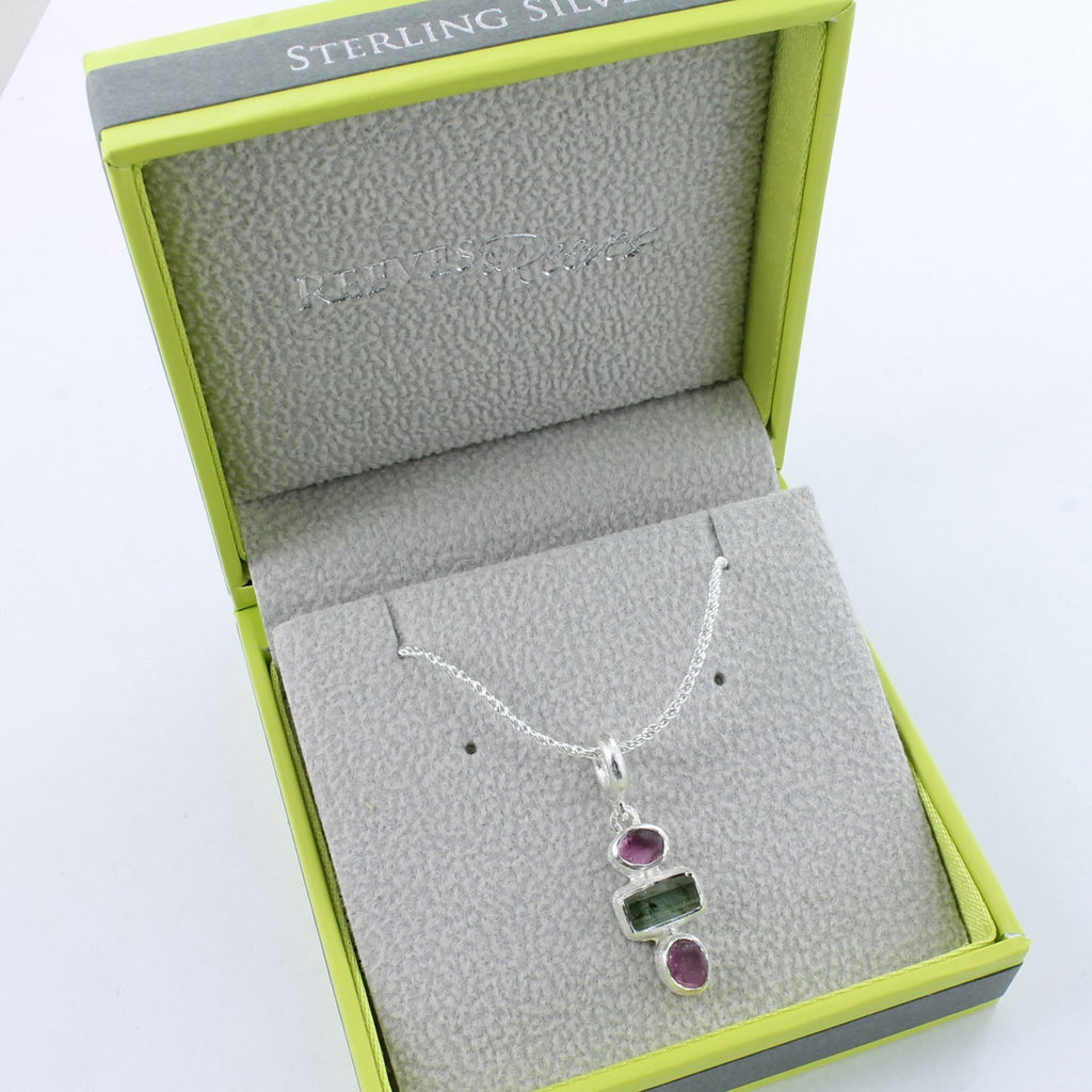 Three stone necklace with two circular rough cut ruby stones and a square peridot sat in the centre. Hung on a silver rope style chain and shown in a Reeves & Reeves gift box.