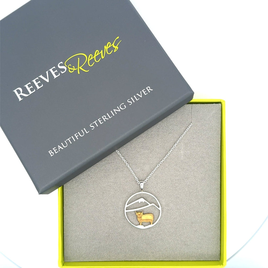 A sterling silver circular pendant with an 18ct gold plated highland cow with silver hills inside the circle. The pendant hangs from a sterling silver adjustable chain and is shown in a Reeves & Reeves gift box.