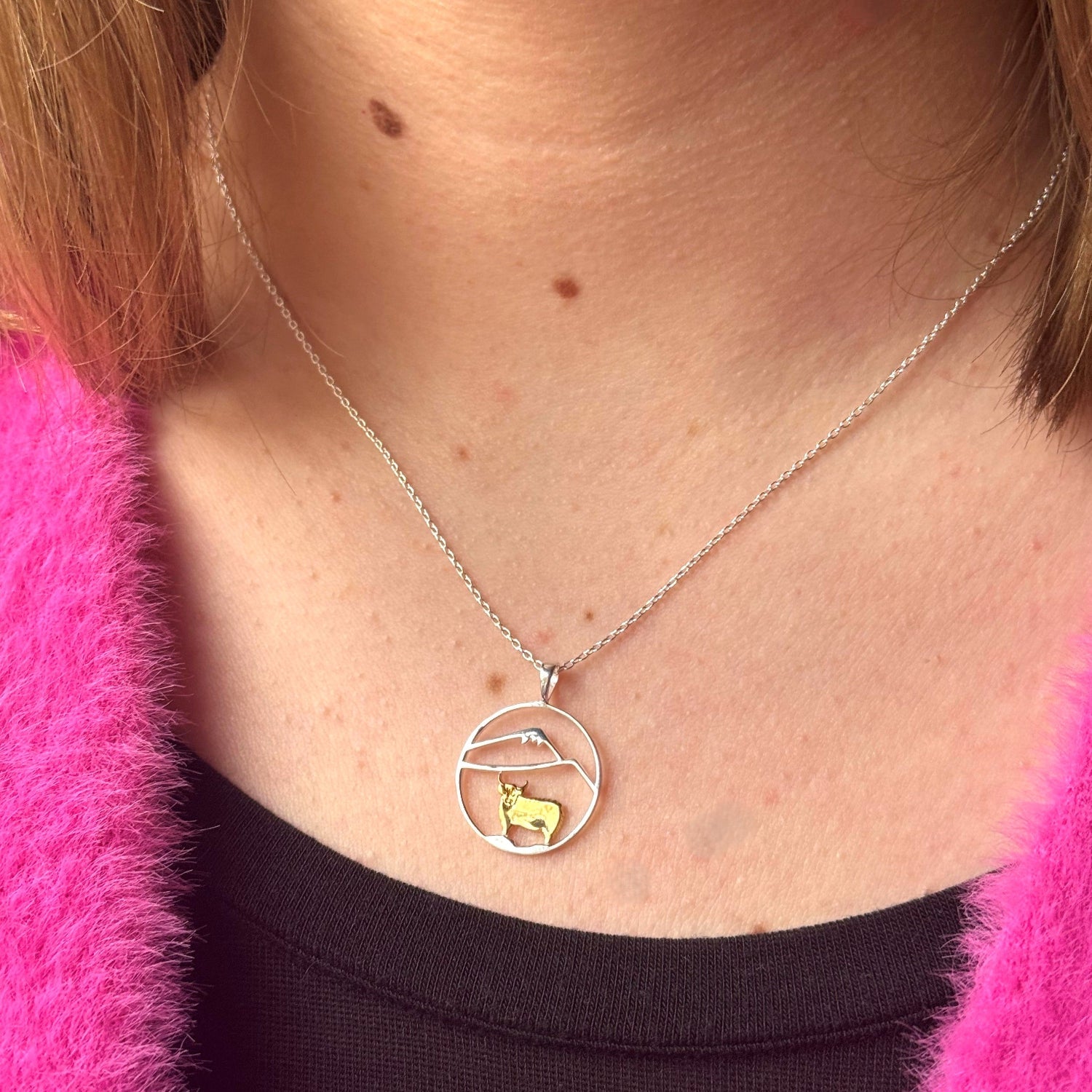 A sterling silver circular pendant with an 18ct gold plated highland cow with silver hills inside the circle. The pendant hangs from a sterling silver adjustable chain and is worn on a model.