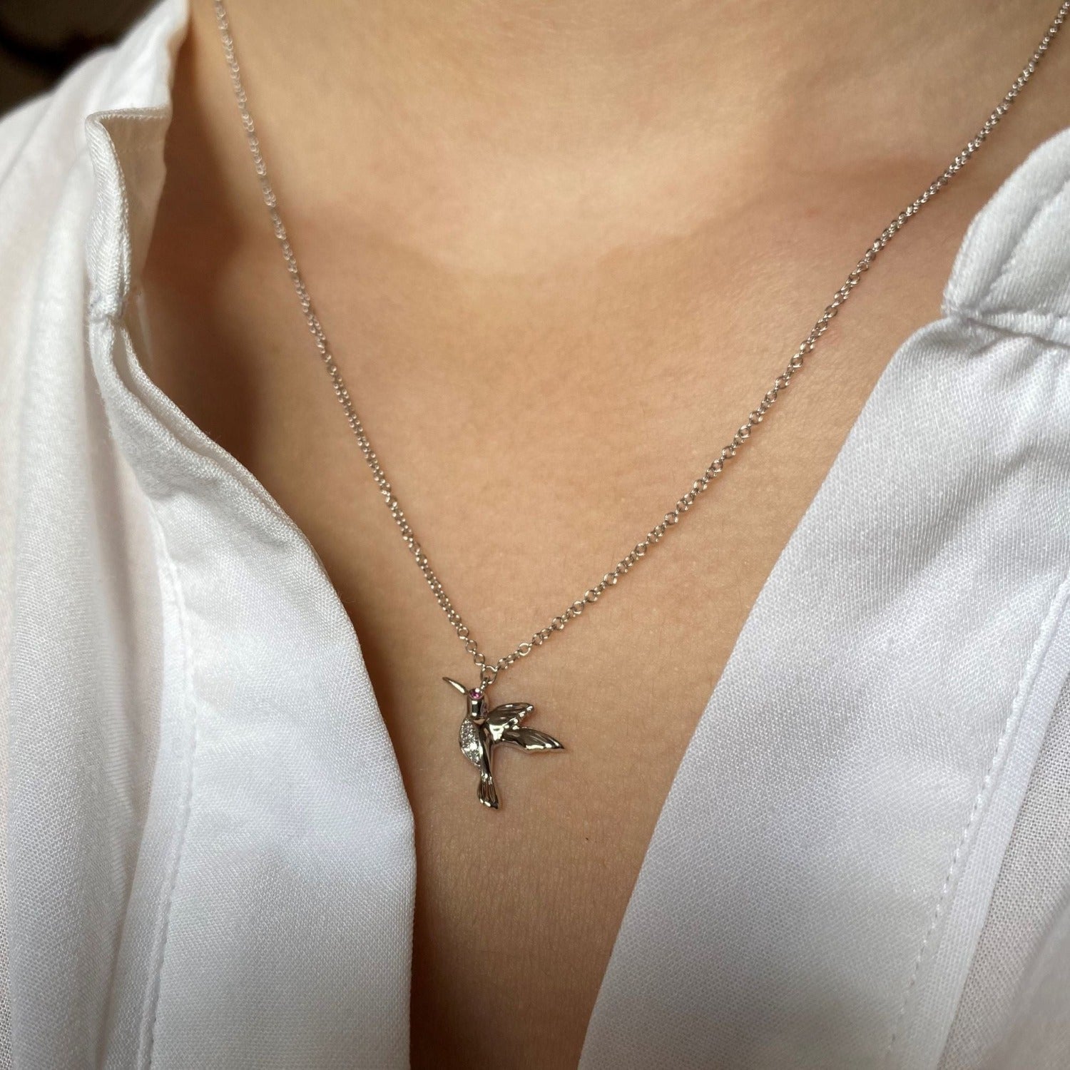 A 14k solid gold hummingbird pendant with diamonds on its tummy and a ruby in its eye. Hung on a solid gold chain and worn on a model.