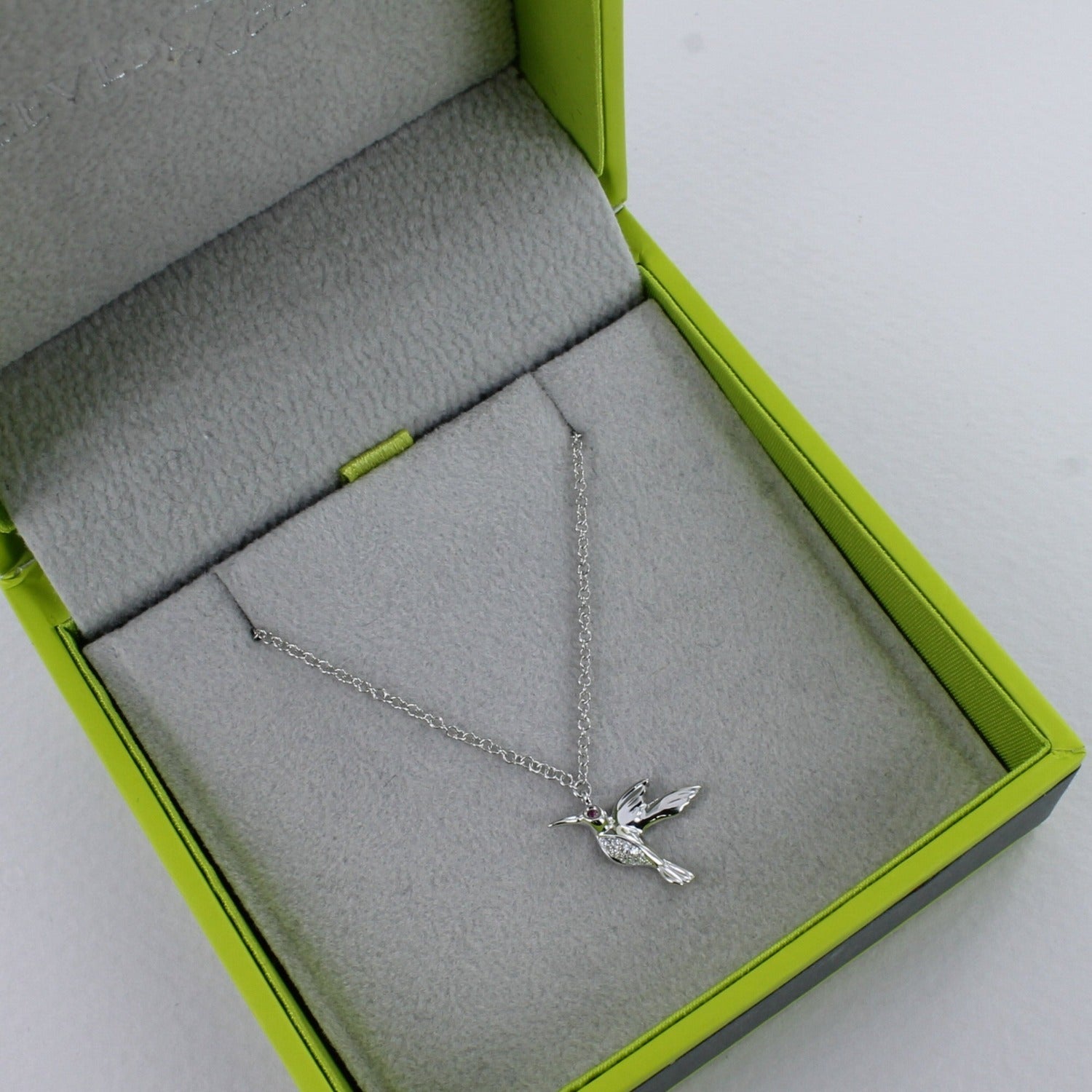A 14k solid white gold hummingbird necklace, with diamonds on its tummy and a ruby in its eye, hung on a solid white gold chain and shown in a Reeves & Reeves gift box.