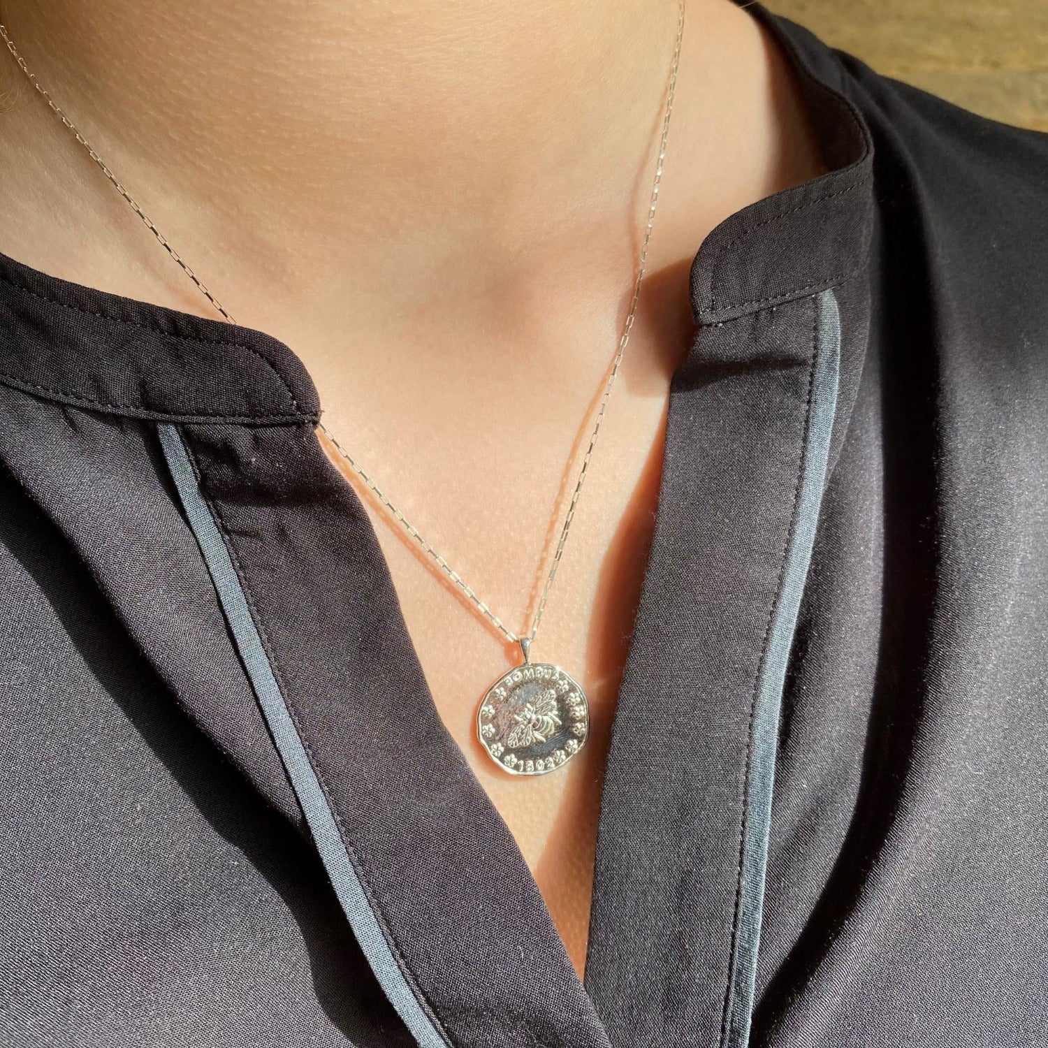 A silver circular pendant with bee detail and bombus written above, hanging on a silver chain and worn on a ladies neck.