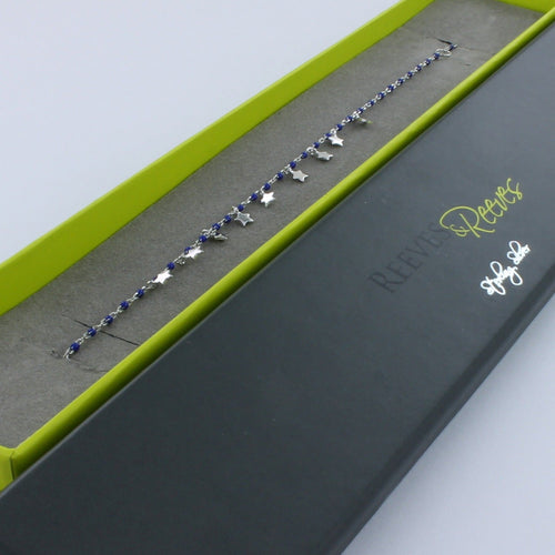 Silver fine chain bracelet with navy blue beads spread evenly along the chain. High shine silver star charms hang from the central part of the chain. Shown in a Reeves & Reeves branded gift box.